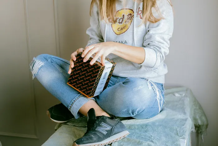 A person sitting on a green covered surface, wearing a gray sweatshirt and blue ripped jeans, opening a black and orange patterned box, possibly containing technology or electronics.