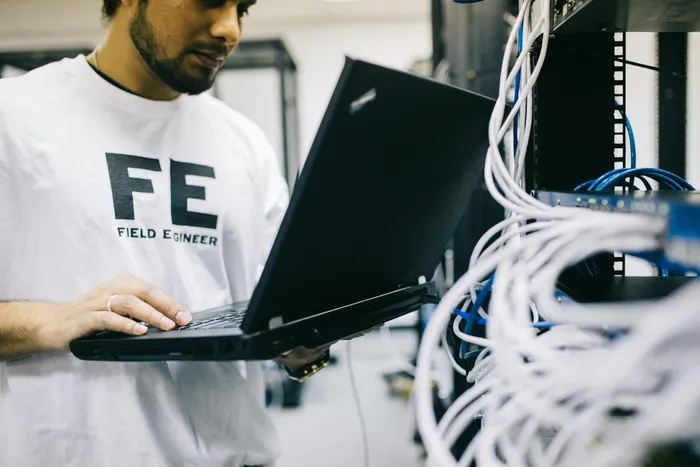 A technician wearing a shirt labeled 'FE' for Field Engineer, standing close to a server rack while working on a laptop, examining network connections and equipment.