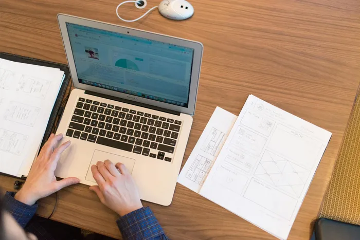 A person's hands typing on a laptop with a view of a WiFi setup guide on the screen, along with printed wireframes and sketches on the table.
